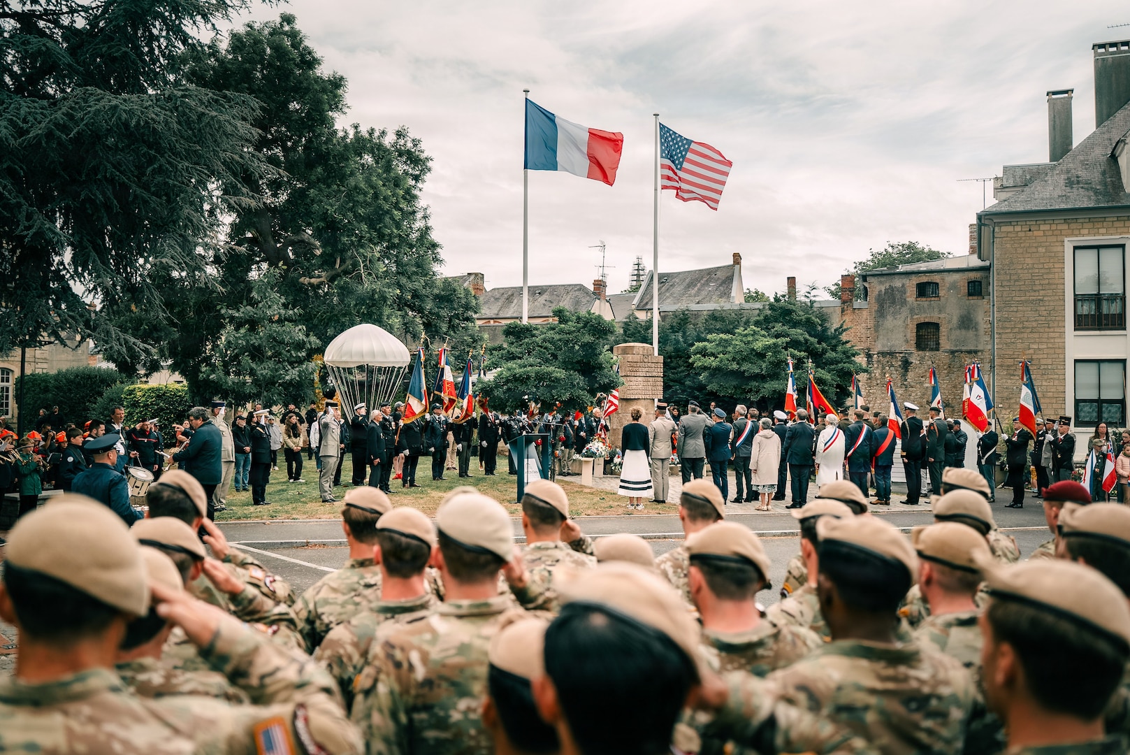 Soldiers stand in formation and salute amid a crowd standing outdoors by a French and a U.S. flag.