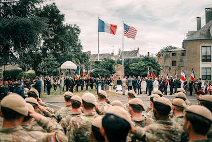 Soldiers stand in formation and salute amid a crowd standing outdoors by a French and a U.S. flag.