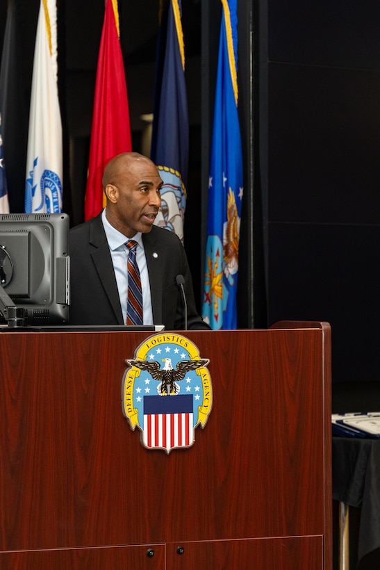 A man wearing a suit stands behind a podium speaking.