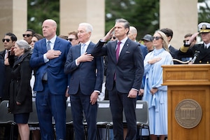 Secretary of War Pete Hegseth salutes as other civilians hold their hands over their hearts while standing outdoors.