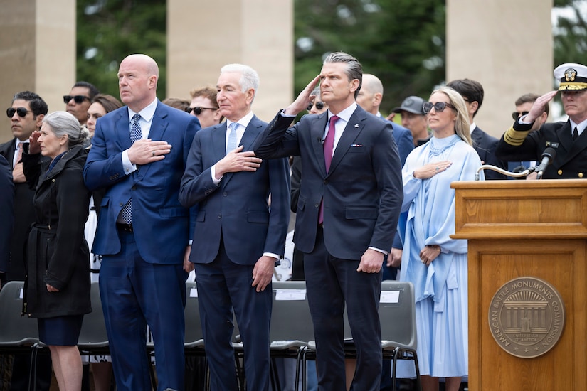 Secretary of War Pete Hegseth salutes as other civilians hold their hands over their hearts while standing outdoors.
