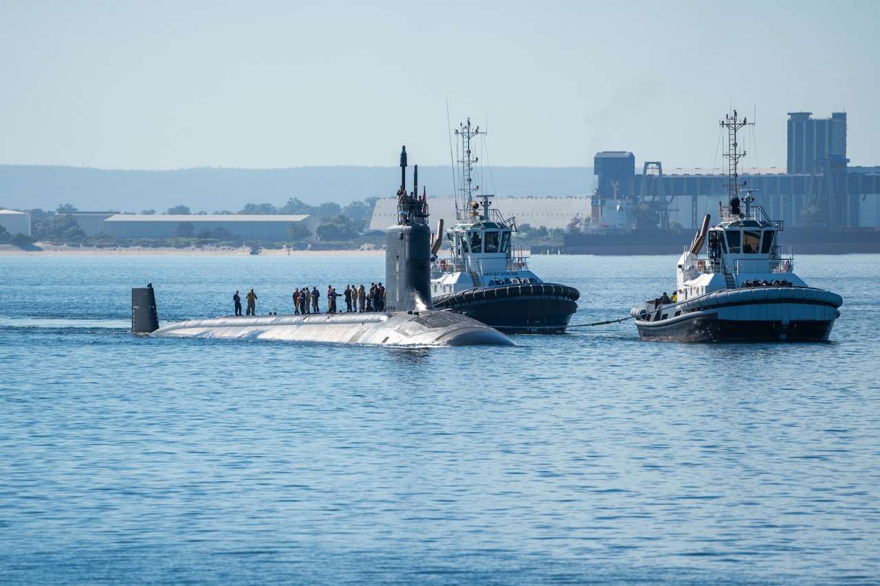 Two tugboats guide a submarine into port while a dozen people stand on top of the sub.