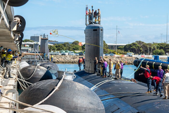 Dozens of people in military uniforms stand on top of a submarine as one person throws a rope to another person standing on a pier.