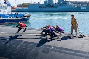 Five men in military uniforms and brightly colored vests attach mooring lines to a submarine in a bay.