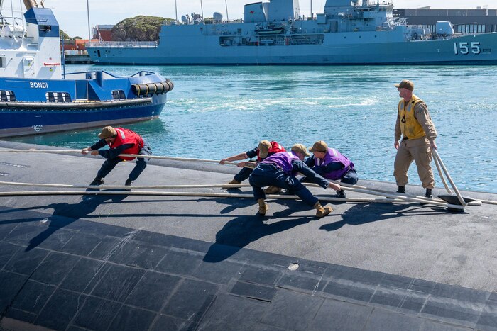 Five men in military uniforms and brightly colored vests attach mooring lines to a submarine in a bay.
