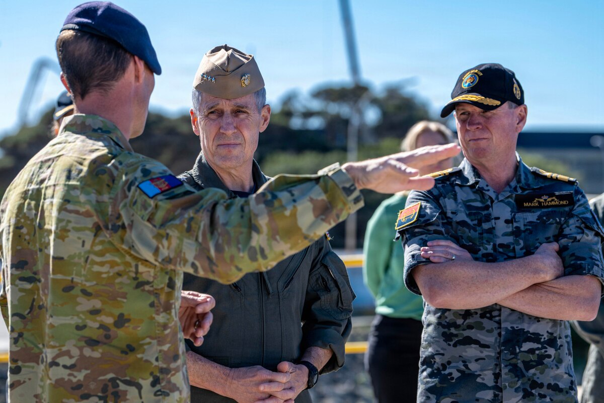 Two men in military uniforms listen to another man in a military camouflage uniform as he speaks to them while standing on a pier.