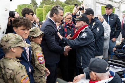 Secretary of War Pete Hegseth shakes hands with a veteran in an outdoor space amid a crowd of adults and kids.