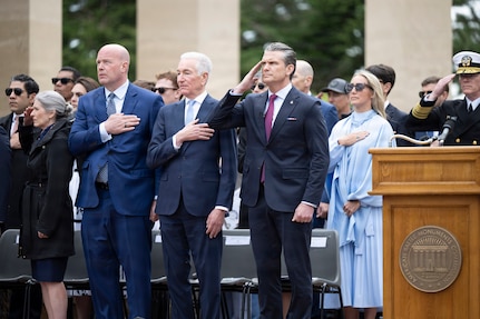 Secretary of War Pete Hegseth salutes as other civilians hold their hands over their hearts while standing outdoors.