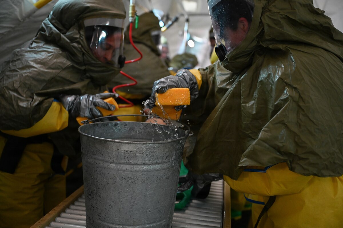Two people wearing hazmat gear rinse out a steel bucket with a red water hose while scrubbing it with sponges.