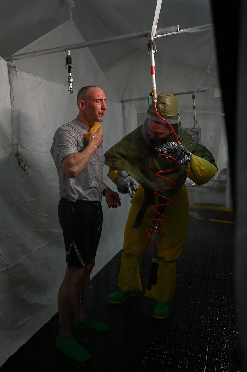 A man wearing hazmat gear rinses off while another man wearing athletic gear standing next to him does the same.
