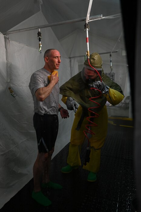 A man wearing hazmat gear rinses off while another man wearing athletic gear standing next to him does the same.
