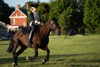 Someone dressed in a dark blue Revolutionary War-era military uniform is on a horse performing the role of George Washington during an outdoor event.