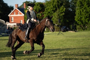 Someone dressed in a dark blue Revolutionary War-era military uniform is on a horse performing the role of George Washington during an outdoor event.