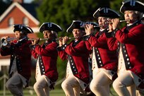 Army soldiers dressed in red Revolutionary War-era uniforms are performing on fifes and bugles while marching during an outdoor event.