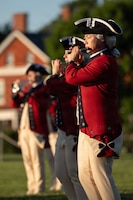 Army soldiers dressed in red Revolutionary War-era uniforms are performing on fifes during an outdoor event.