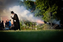 Black cannons with the words U.S. Army on barrels are firing blanks during an outdoor show. In the foreground, someone dressed in a long black coat and black top hat is walking across the field.