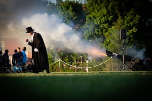 Black cannons with the words U.S. Army on barrels are firing blanks during an outdoor show. In the foreground, someone dressed in a long black coat and black top hat is walking across the field.