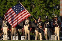 Army soldiers dressed in early 1800s-era military uniforms are standing in a formation during an outdoor show while one soldier waves a period U.S. flag. The other soldiers are holding rifles with bayonets affixed.