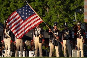 Army soldiers dressed in early 1800s-era military uniforms are standing in a formation during an outdoor show while one soldier waves a period U.S. flag. The other soldiers are holding rifles with bayonets affixed.