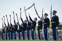 Army soldiers dressed in dark blue ceremonial uniforms are standing in a row while tossing rifles into the air during an outdoor show. Audience members are seated on bleachers in the background.