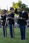 Army soldiers dressed in dark blue ceremonial uniforms are standing in a row performing a drill routine. While the other are holding their rifles in front of their bodies, one soldier is flexing his bicep at the audience for this outdoor show.