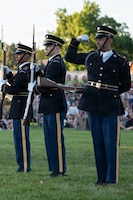 Army soldiers dressed in dark blue ceremonial uniforms are standing in a row performing a drill routine. While the other are holding their rifles in front of their bodies, one soldier is flexing his bicep at the audience for this outdoor show.