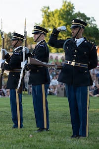 Army soldiers dressed in dark blue ceremonial uniforms are standing in a row performing a drill routine. While the other are holding their rifles in front of their bodies, one soldier is flexing his bicep at the audience for this outdoor show.