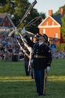 Army soldiers wearing dark blue ceremonial uniforms are standing in a straight row while tossing rifles into the air. There are hundreds of people sitting on bleachers in the background watching the outdoor show.