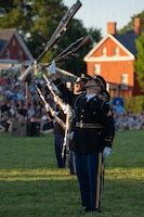 Army soldiers wearing dark blue ceremonial uniforms are standing in a straight row while tossing rifles into the air. There are hundreds of people sitting on bleachers in the background watching the outdoor show.