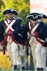 Army soldiers dressed in Revolutionary War-era uniforms are charging holding rifles with bayonets affixed during a demonstration at an outdoor show.