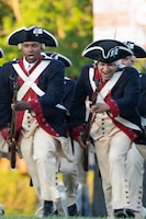 Army soldiers dressed in Revolutionary War-era uniforms are charging holding rifles with bayonets affixed during a demonstration at an outdoor show.