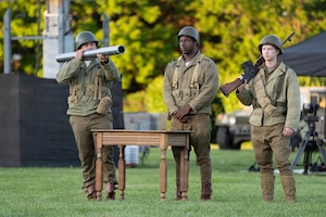 Three Army soldiers wearing historic green Army uniforms with World War 2-era helmets are performing a dramatic vignette during an outdoor show. One is holding a bazooka, and another had a rifle on his shoulder.