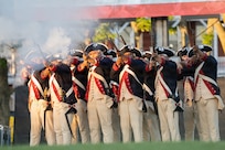 Army Soldiers dressed in Revolutionary War-era uniforms are firing from ceremonial rifles during a demonstration at an outdoor show.