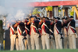 Army Soldiers dressed in Revolutionary War-era uniforms are firing from ceremonial rifles during a demonstration at an outdoor show.