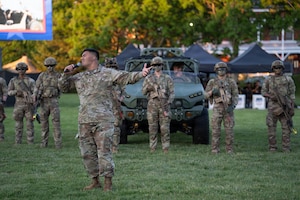 An Army Soldier dressed in combat uniform is holding a microphone while singing at an outdoor event. There are several soldier dressed similarly with helmets on and carrying rifles behind the singer, and their is a large Army vehicle in the background as well.