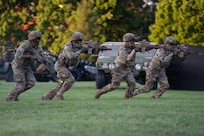 Four Army Soldiers dressed in combat uniforms with helmets and sunglasses on are running across a field while pointing rifles during an outdoor demonstration. There are Army vehicles in the background.