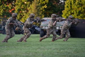 Four Army Soldiers dressed in combat uniforms with helmets and sunglasses on are running across a field while pointing rifles during an outdoor demonstration. There are Army vehicles in the background.