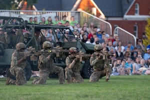 Army soldiers wearing combat uniforms with helmets are aiming rifles in a demonstration at an outdoor show. They are kneeling in front of a green Army vehicle and there are dozens of  people watching from risers in the background.