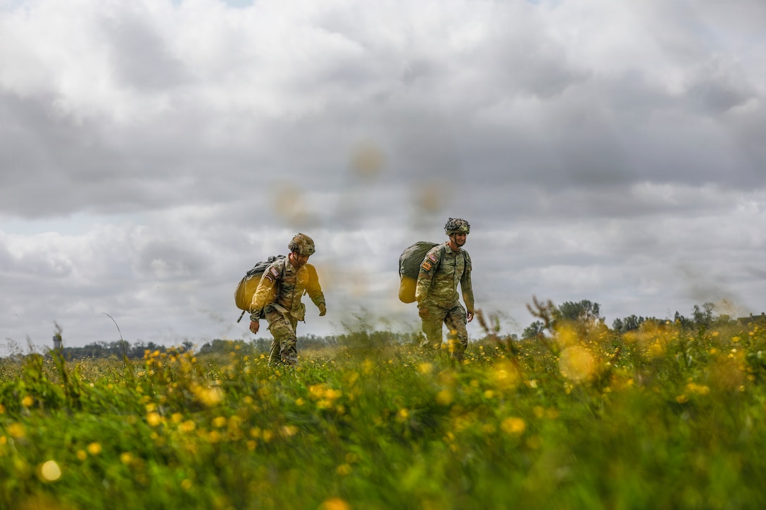 Two soldiers carrying large backpacks walk in a field of yellow flowers under a cloudy sky.