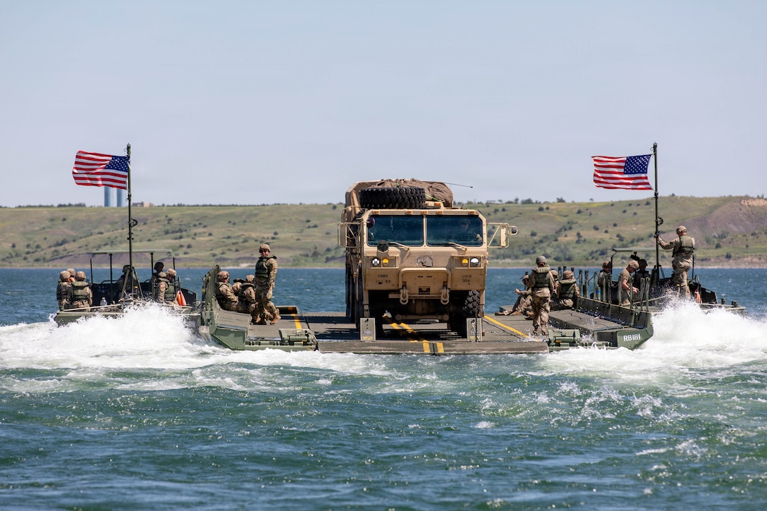 Soldiers in tactical gear ride on an improved ribbon bridge with two American flags to transport a vehicle across a body of water.