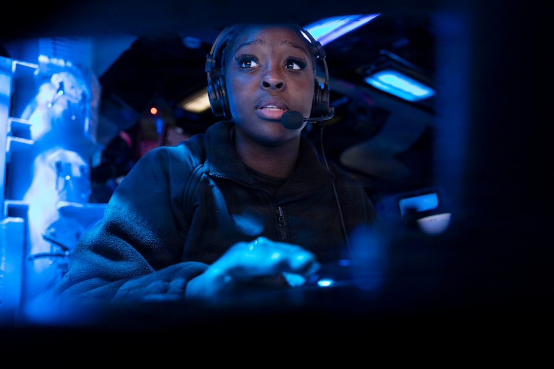 A sailor wearing a headset works in a dark information center illuminated by blue light.