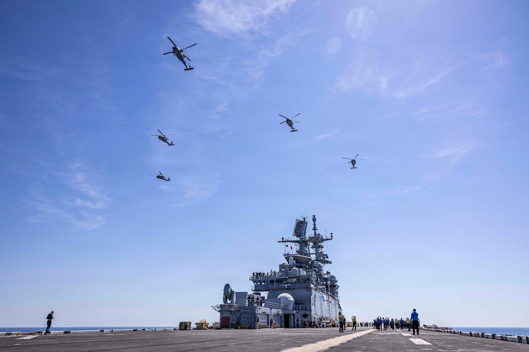 Five helicopters fly in formation over the deck of a ship during the day. Sailors on deck look up.