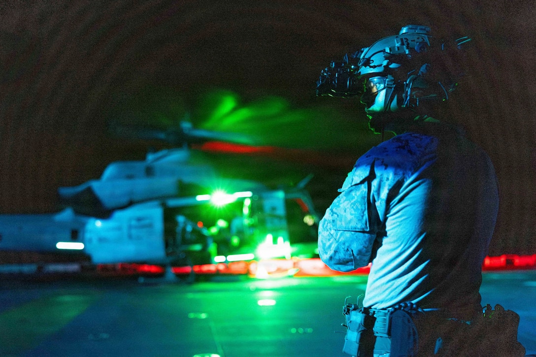 A Marine wearing a helmet with a light attached to it stands on the flight deck of a ship at night. A helicopter with green and red lights is several yards away on the deck.