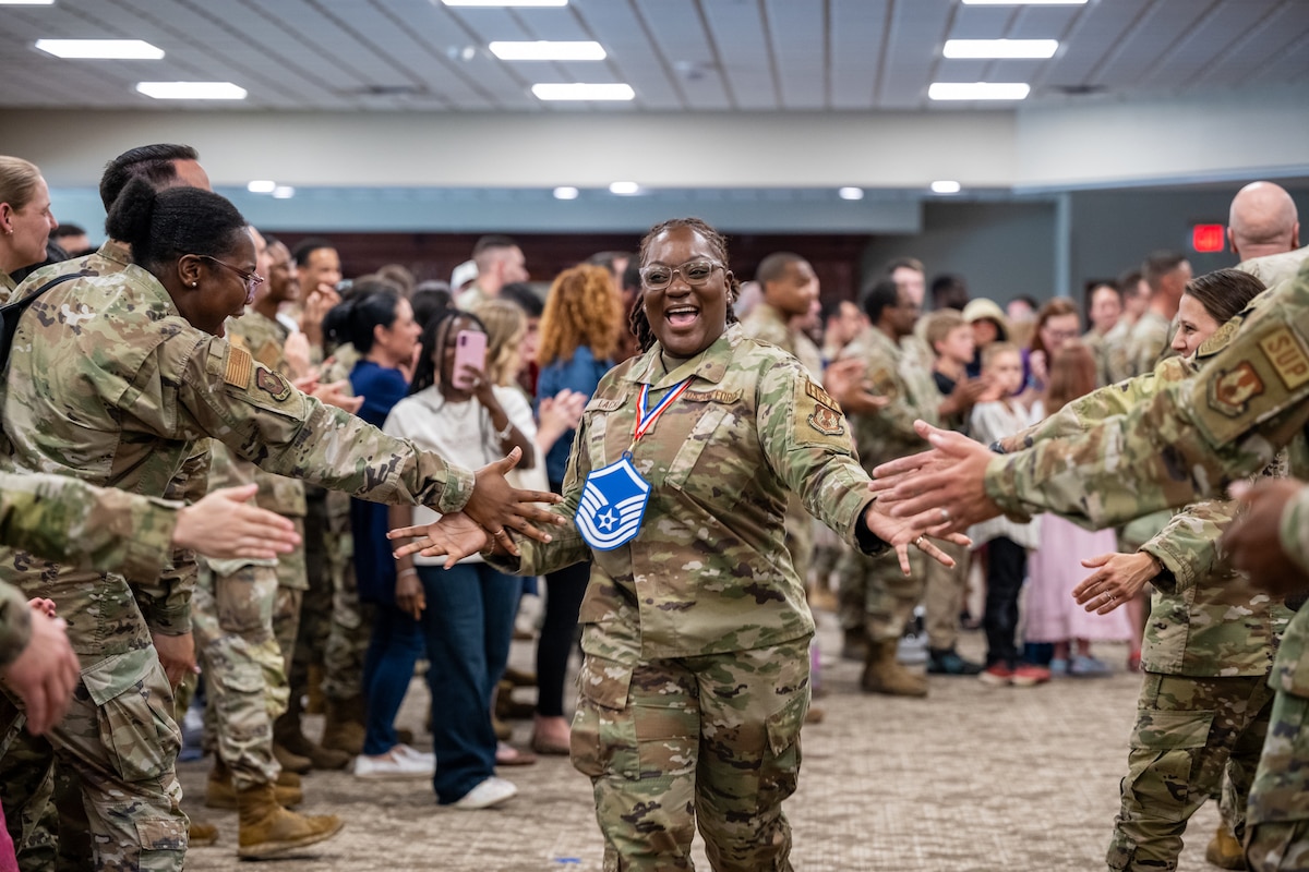 A woman, wearing a military camouflage uniform and master sergeant lanyard, smiles as she is greeted with high fives