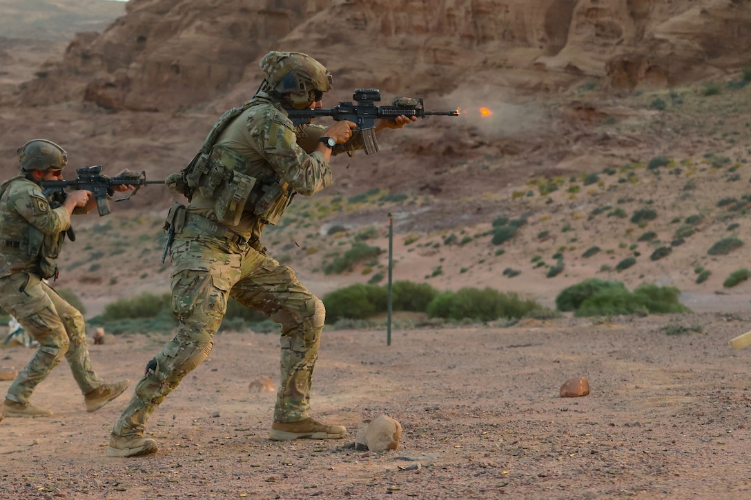 Two soldiers fire rifles in the desert, with mountains in the background.