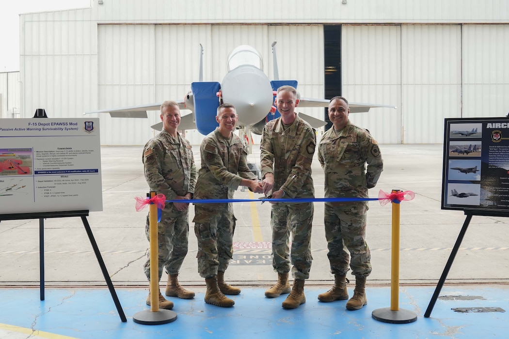 four airmen stand behind a ribbon. two of them in the center are holding a pair of scissors as they prepare to cut the ribbon in front of an F-15 aircraft