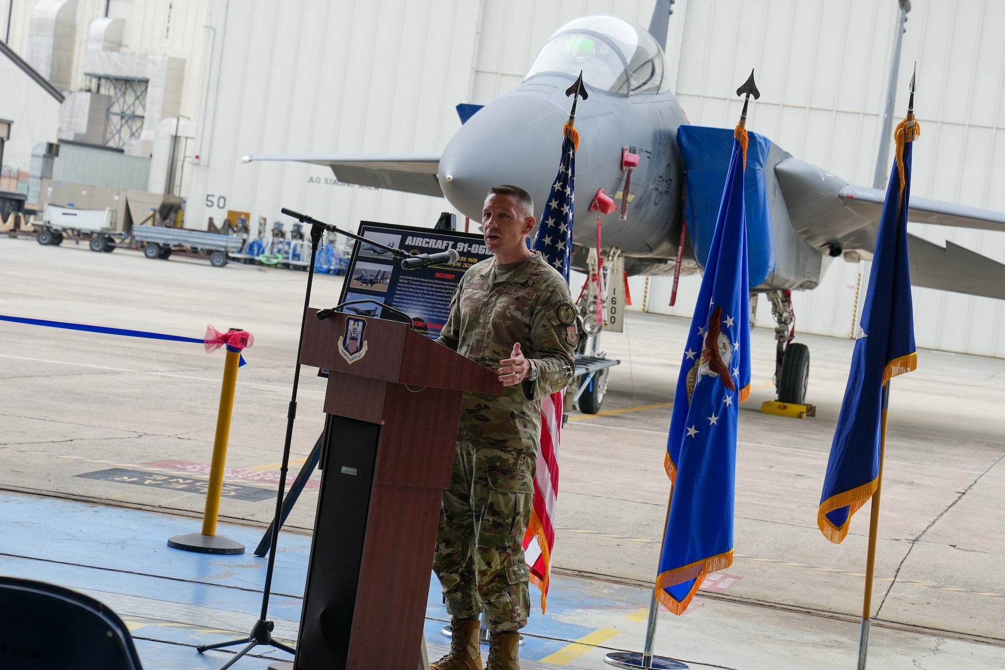 an airman speaks from a podium with three flags and an f-15 in the background behind him
