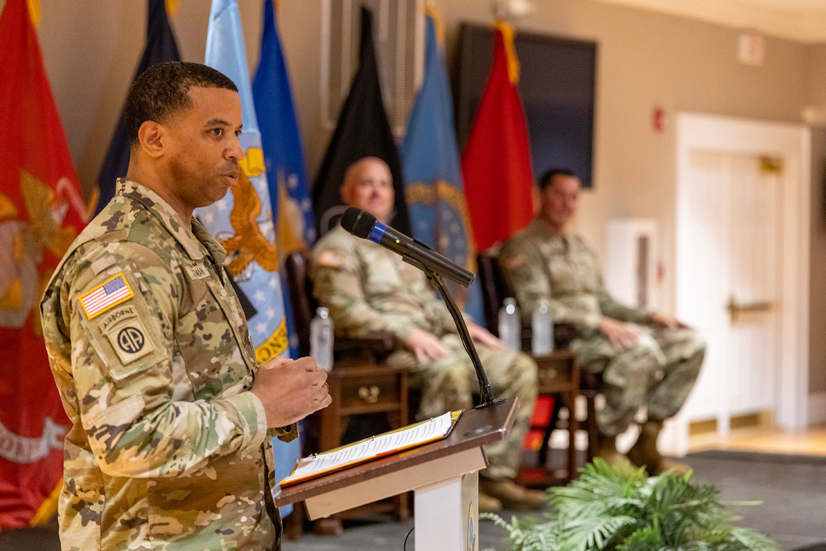 A man wearing a camouflage military uniform stand on stage behind a podium speaking to a seated audience.