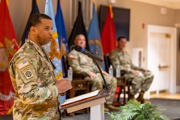 A man wearing a camouflage military uniform stand on stage behind a podium speaking to a seated audience.