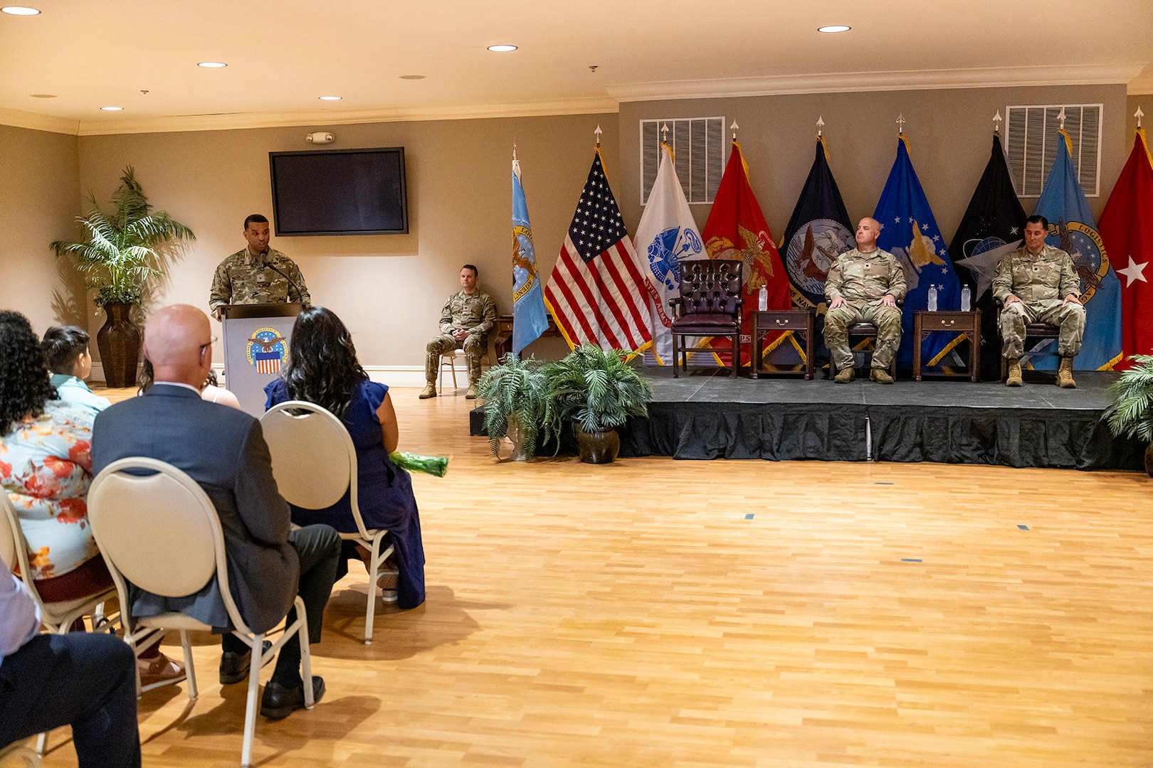 A man wearing a camouflage military uniform stand on stage behind a podium speaking to a seated audience.